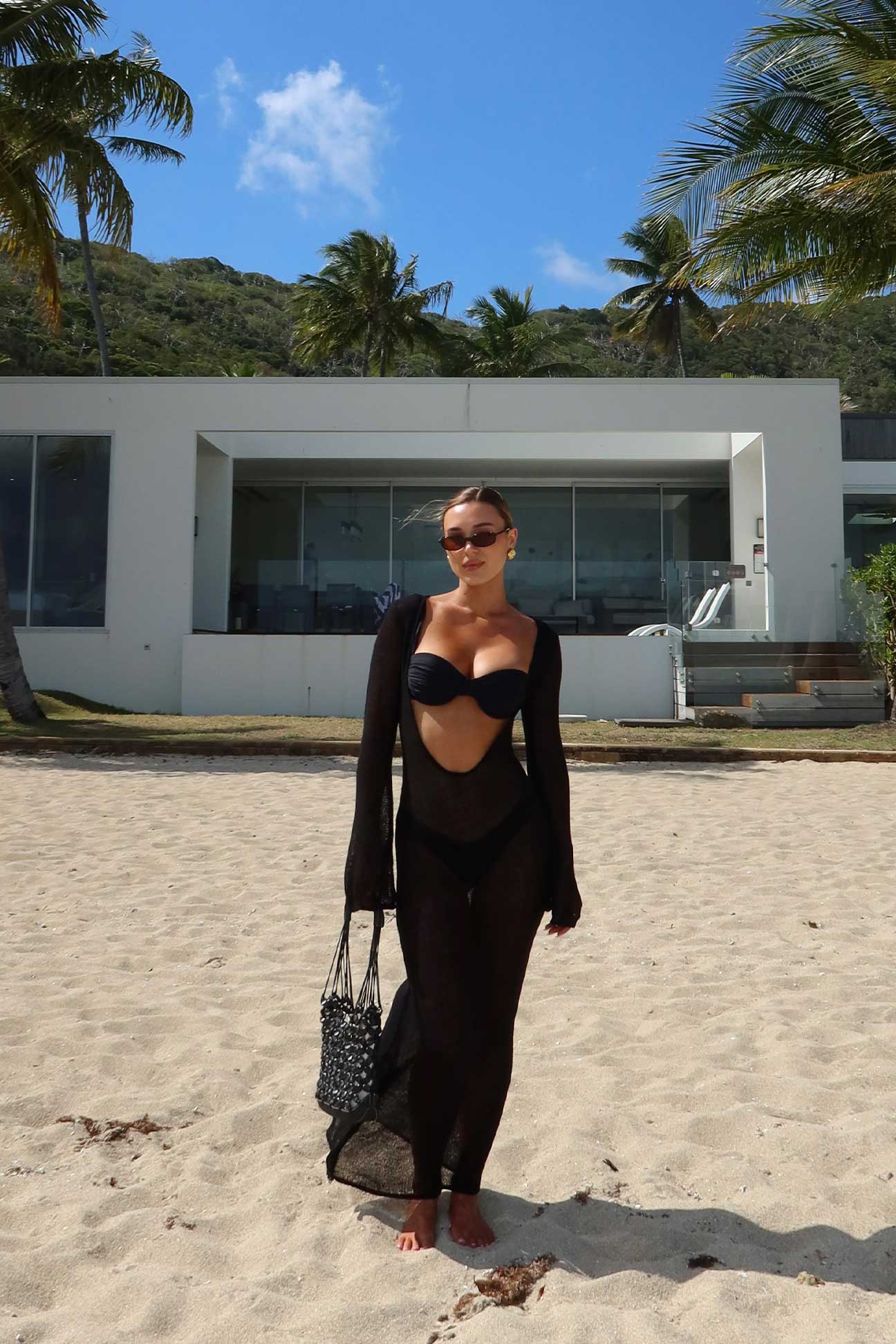 Woman in black dress standing on a sandy beach with palm trees and a building in the background