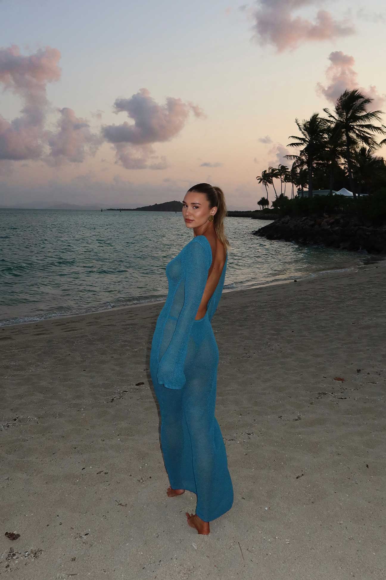 Woman in a blue dress standing on a beach with palm trees and water in the background