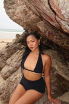 Woman in a black bikini sitting against a rocky beach backdrop
