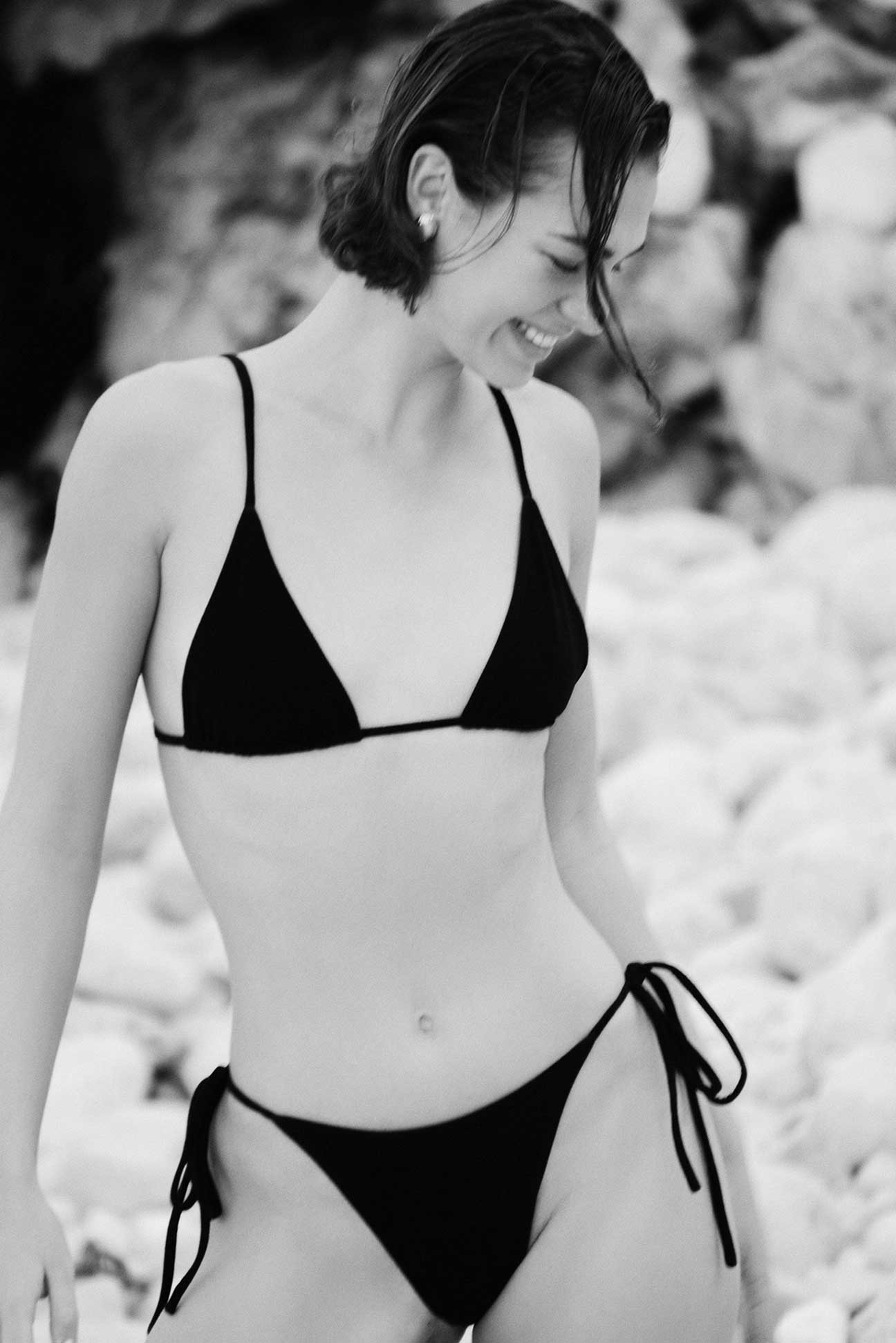 Woman wearing a black bikini standing on a rocky beach.
