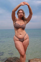 Woman in a mocha bikini standing on rocks by the ocean with a clear sky.