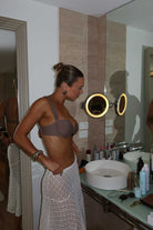 Woman in a bathroom wearing a mocha bikini, standing in front of a mirror.