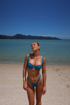Woman in a teal bikini standing on a beach with clear blue water and mountains in the background
