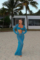 Woman in a teal outfit standing on a sandy beach with palm trees and a building in the background.