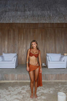Woman in a terracotta bikini standing in a rustic indoor setting with wooden walls and white furniture.