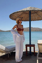 Woman in a terracotta bikini and white cover-up standing under a straw umbrella by the water.