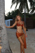 Woman in a terracotta bikini and rust sarong standing by a palm tree on a beach.