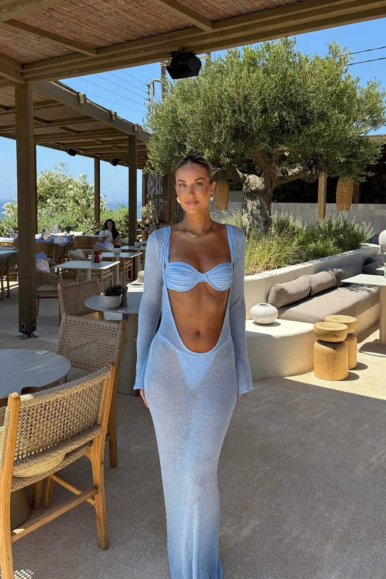 Woman in a light blue dress standing outdoors in a sunlit area with trees and furniture.