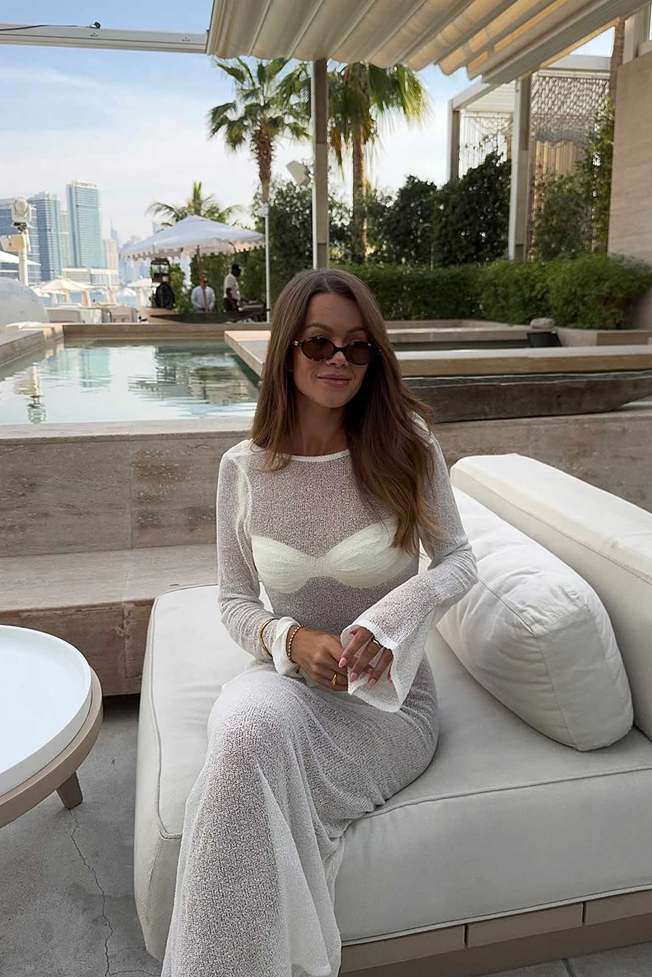 Woman sitting on a white outdoor sofa in a white dress with a pool and cityscape in the background