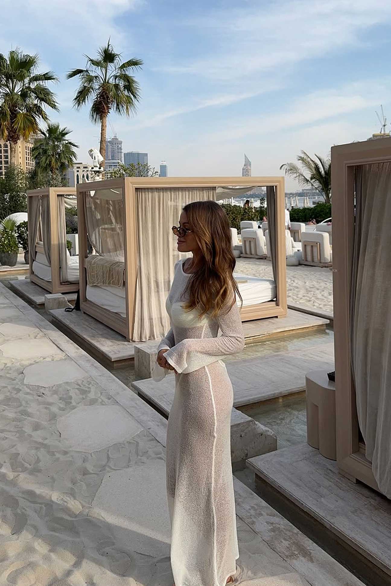Woman in a long, light-colored dress standing by a poolside with palm trees and buildings in the background.
