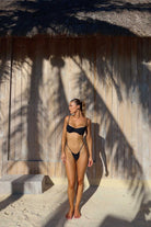 Woman in a black bikini standing under a thatched roof with palm shadows.