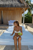 Woman in a bikini sitting on an inflatable bed with a thatched-roof structure and palm trees in the background.