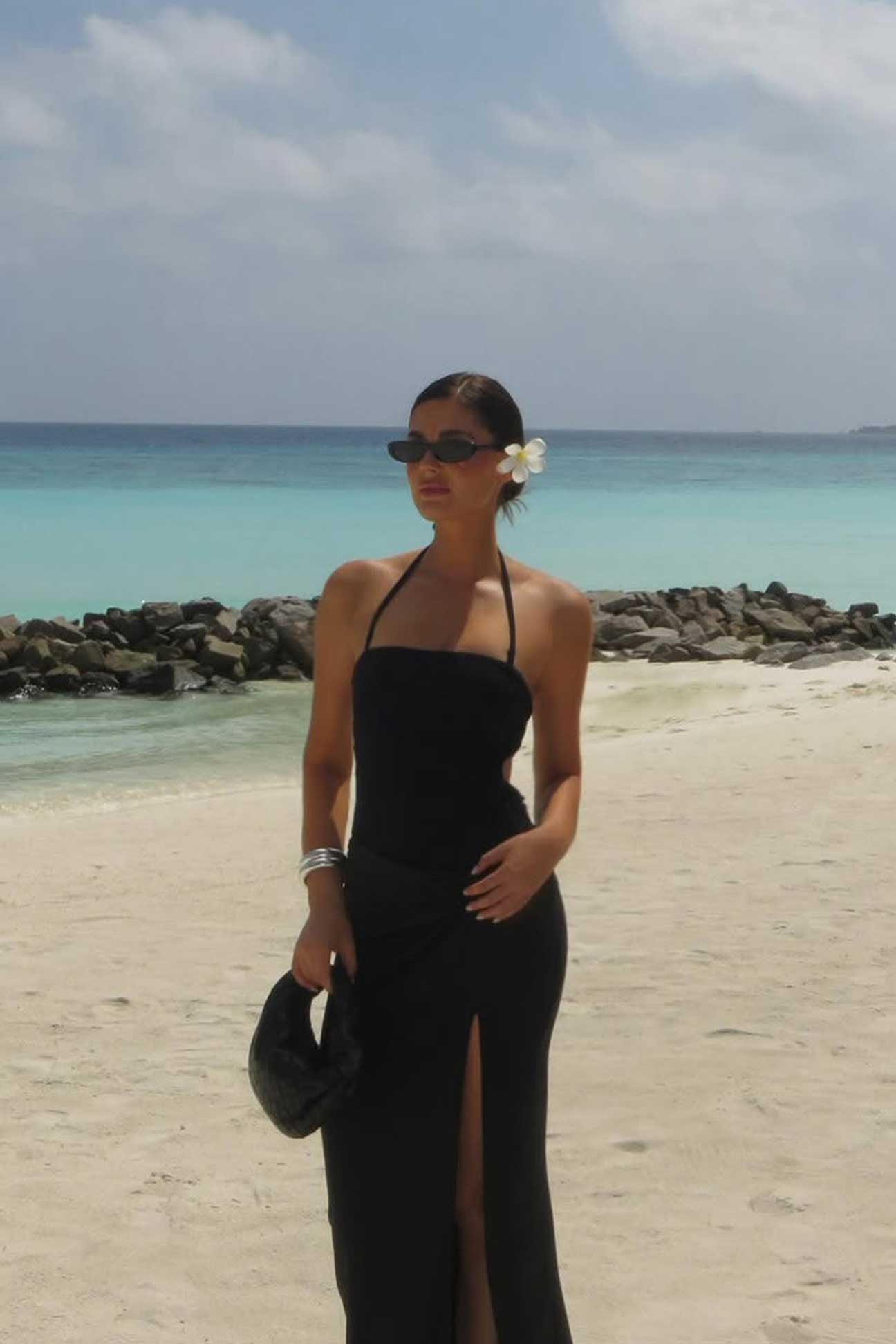 Woman in a black dress standing on a beach with clear blue water and sky.