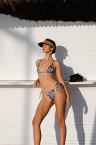 Woman in a floral bikini and hat standing against a white wall with a thatched roof structure.