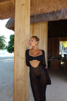Woman in a black outfit standing under a thatched-roof structure with a beach setting.