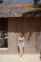 Woman in a white outfit standing in front of a wooden wall with a thatched roof structure.
