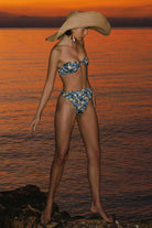 Woman in a floral bikini and large hat standing on rocks by the water at sunset.