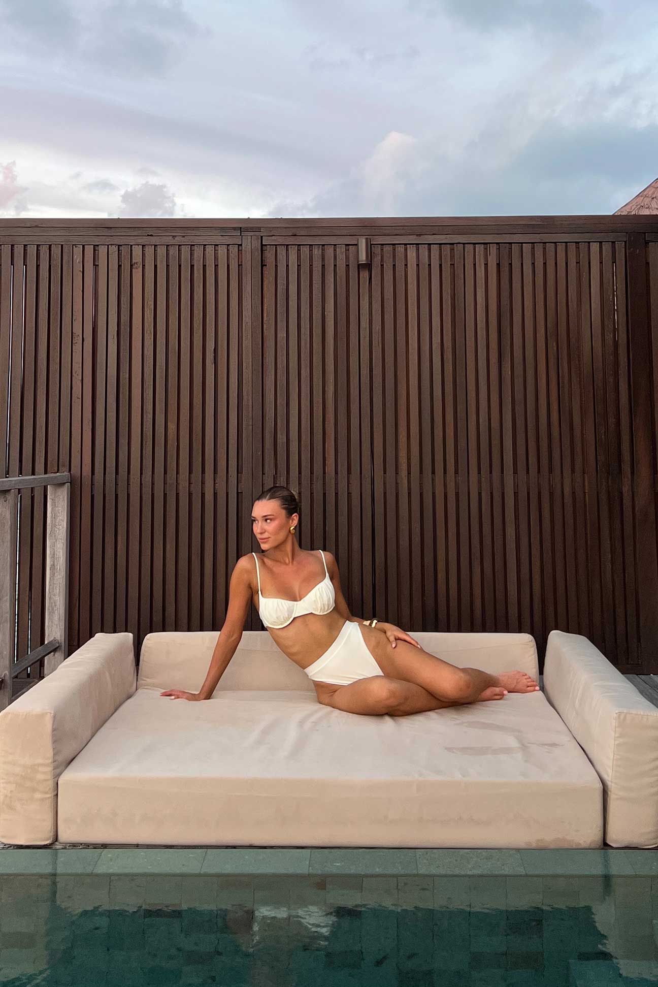 Woman in a bikini sitting on a floating platform by a pool with a wooden wall in the background.