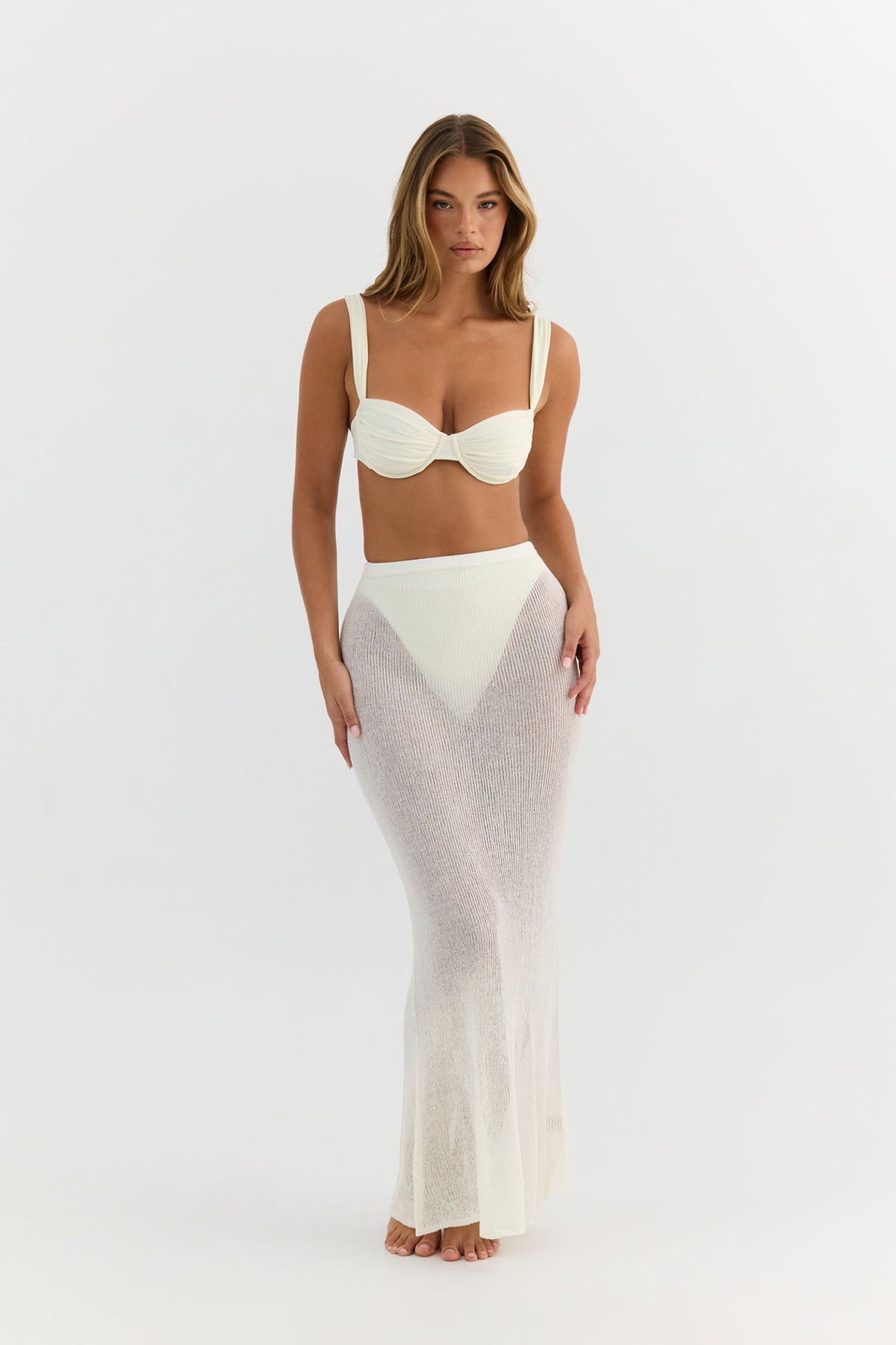A woman modeling a two-piece white outfit consisting of a structured bralette top and a sheer, textured maxi skirt, photographed against a seamless white studio background.