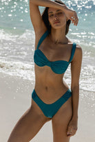 Woman in a teal bikini standing on a beach with ocean in the background