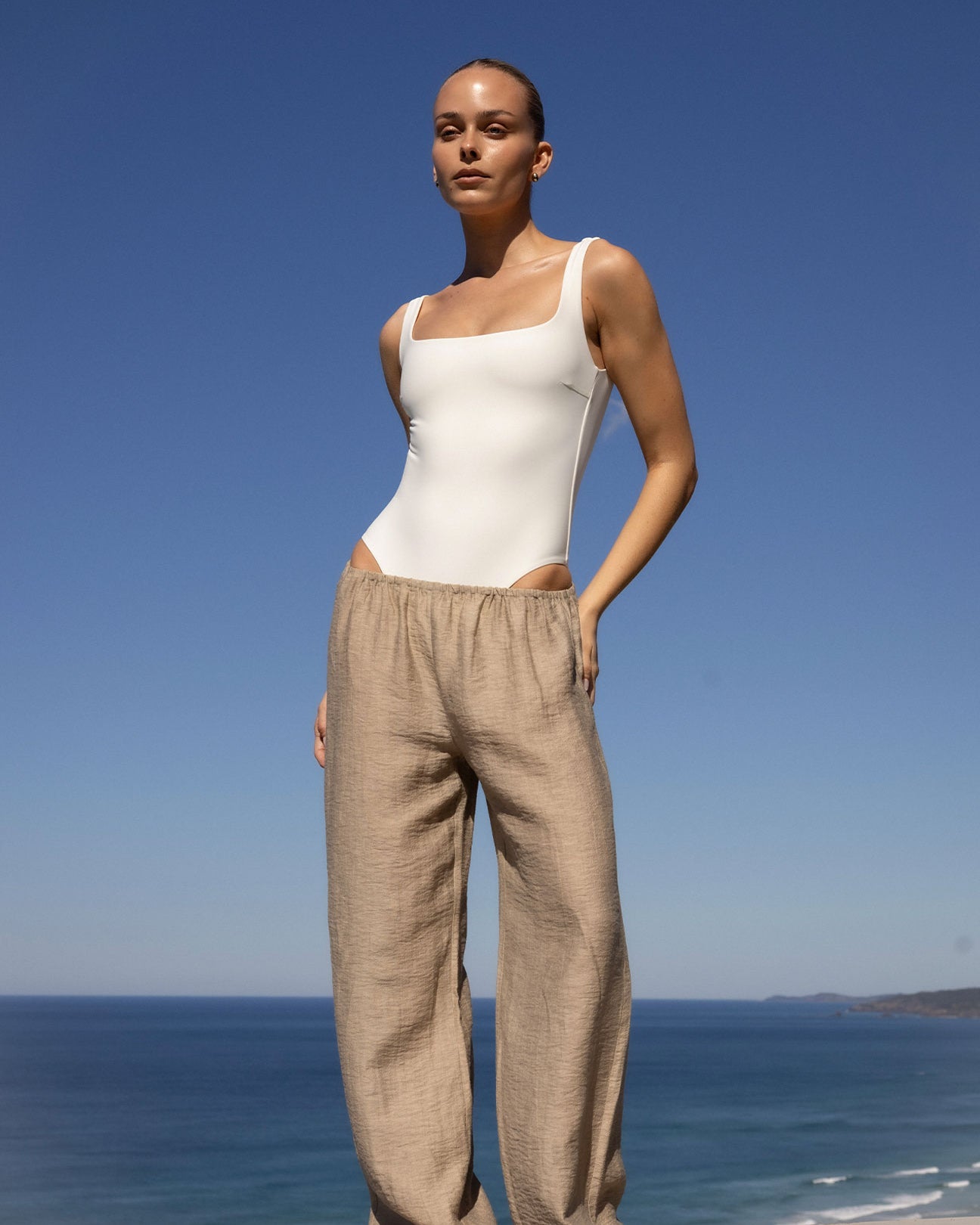 A woman stands outdoors by the ocean wearing a fitted white bodysuit and loose beige linen pants against a clear blue sky.