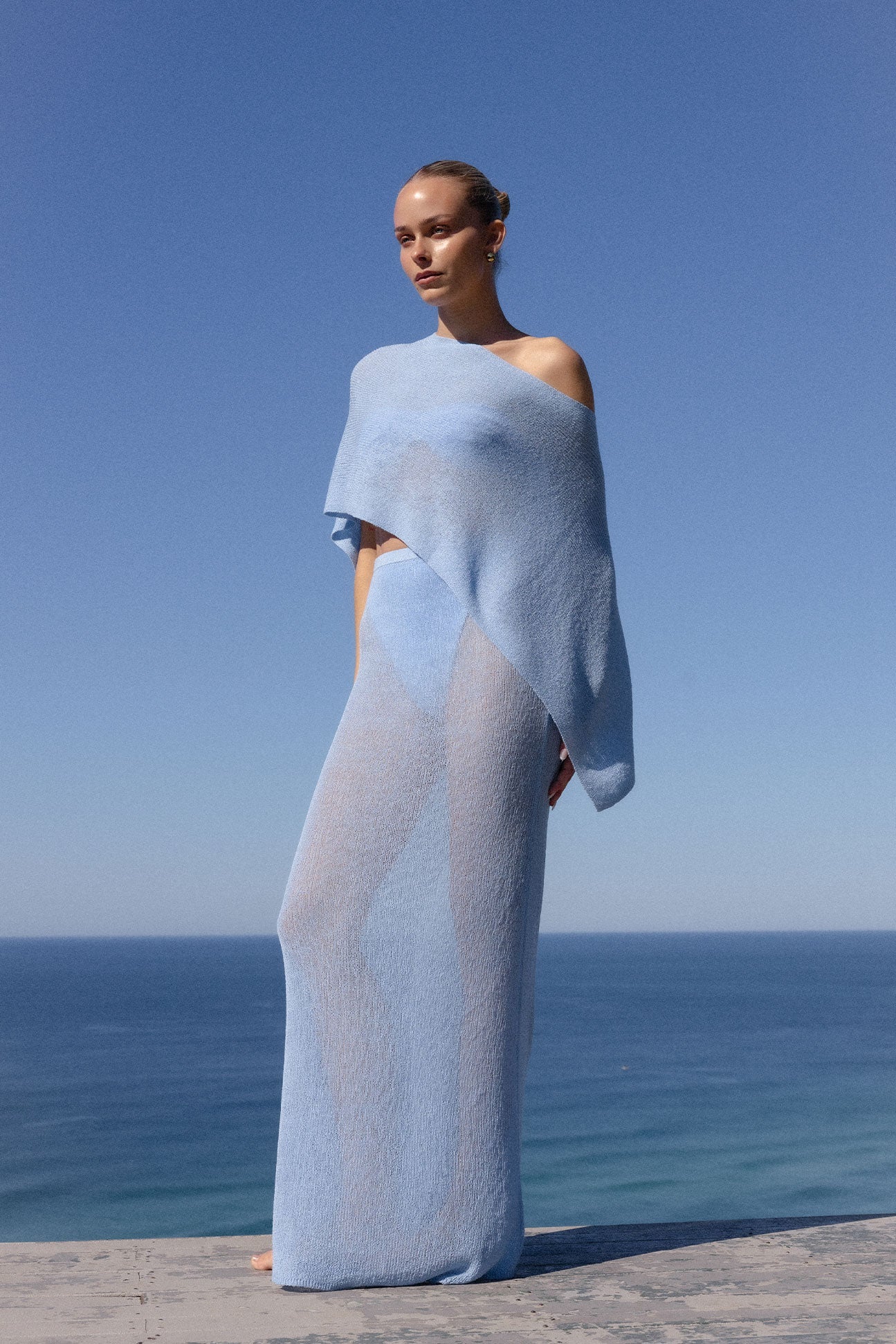 A woman stands outdoors by the sea wearing a flowing, semi-sheer light blue wrap dress or cover-up with draped sleeves against a clear blue sky.