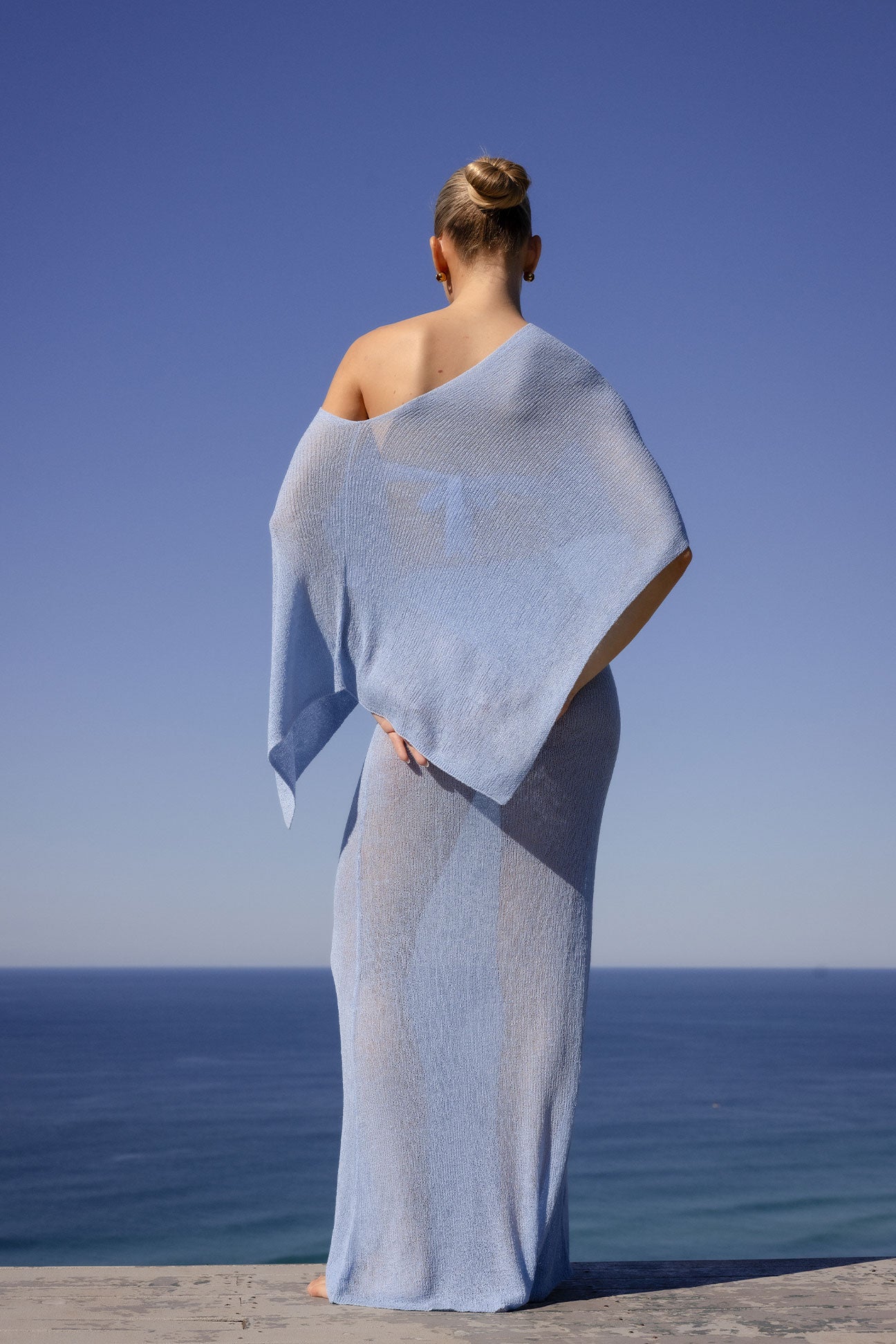 A woman stands with her back to the camera wearing a light blue sheer knit wrap that drapes over her body, set against a clear blue sky and ocean background.