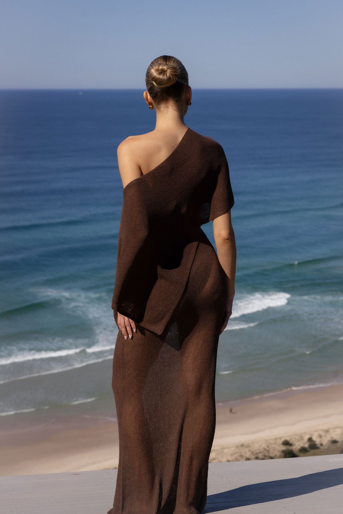 A woman stands with her back to the camera, wearing a long, dark brown off-the-shoulder knit dress, overlooking a beach and the blue ocean.