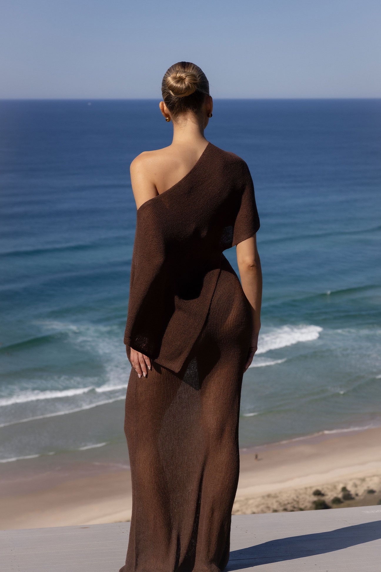 A woman stands with her back to the camera, wearing a long, dark brown off-the-shoulder knit dress, overlooking a beach and the blue ocean.