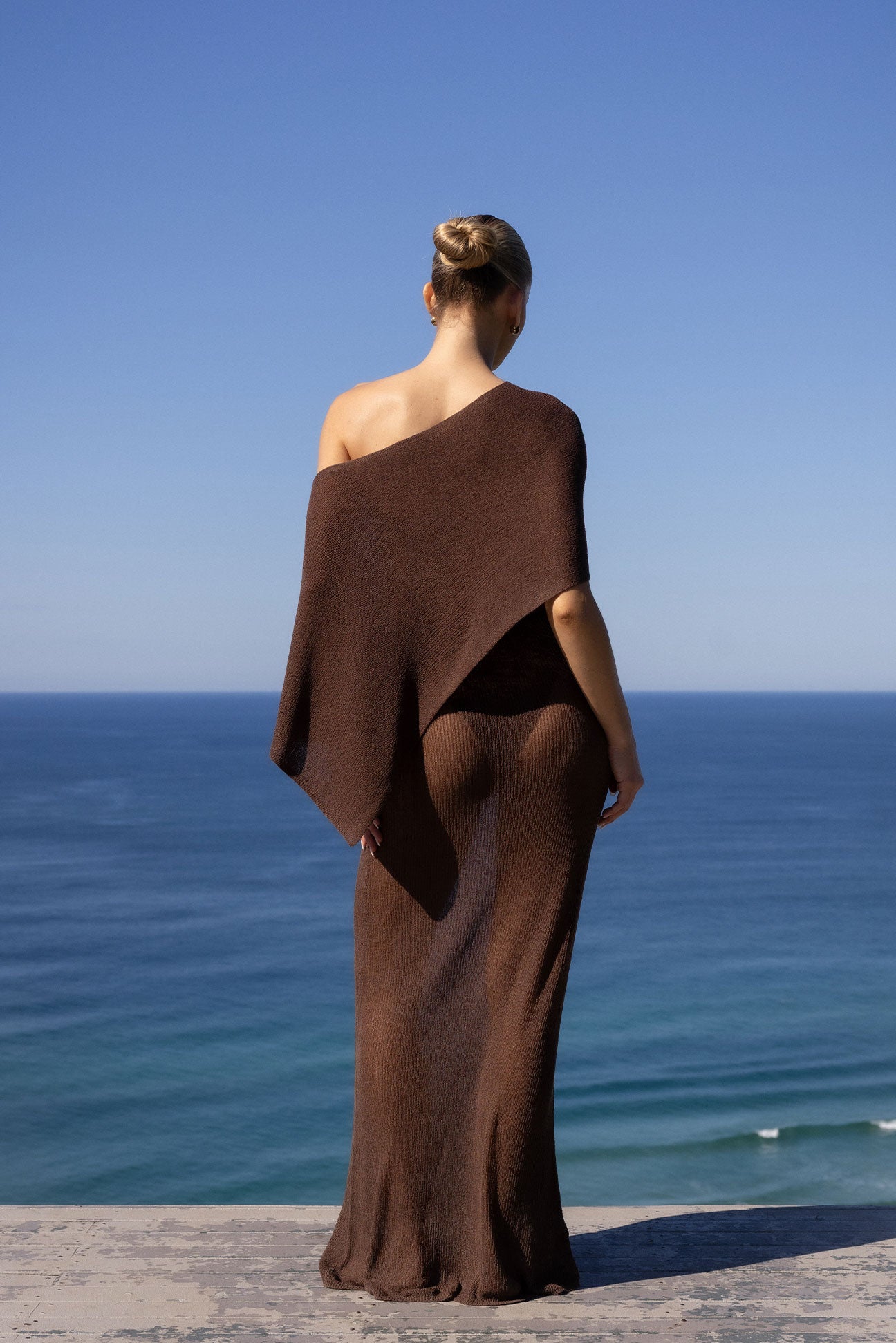 A woman stands with her back to the camera wearing a long, draped brown one-shoulder wrap dress or shawl, overlooking a blue ocean under a clear sky.
