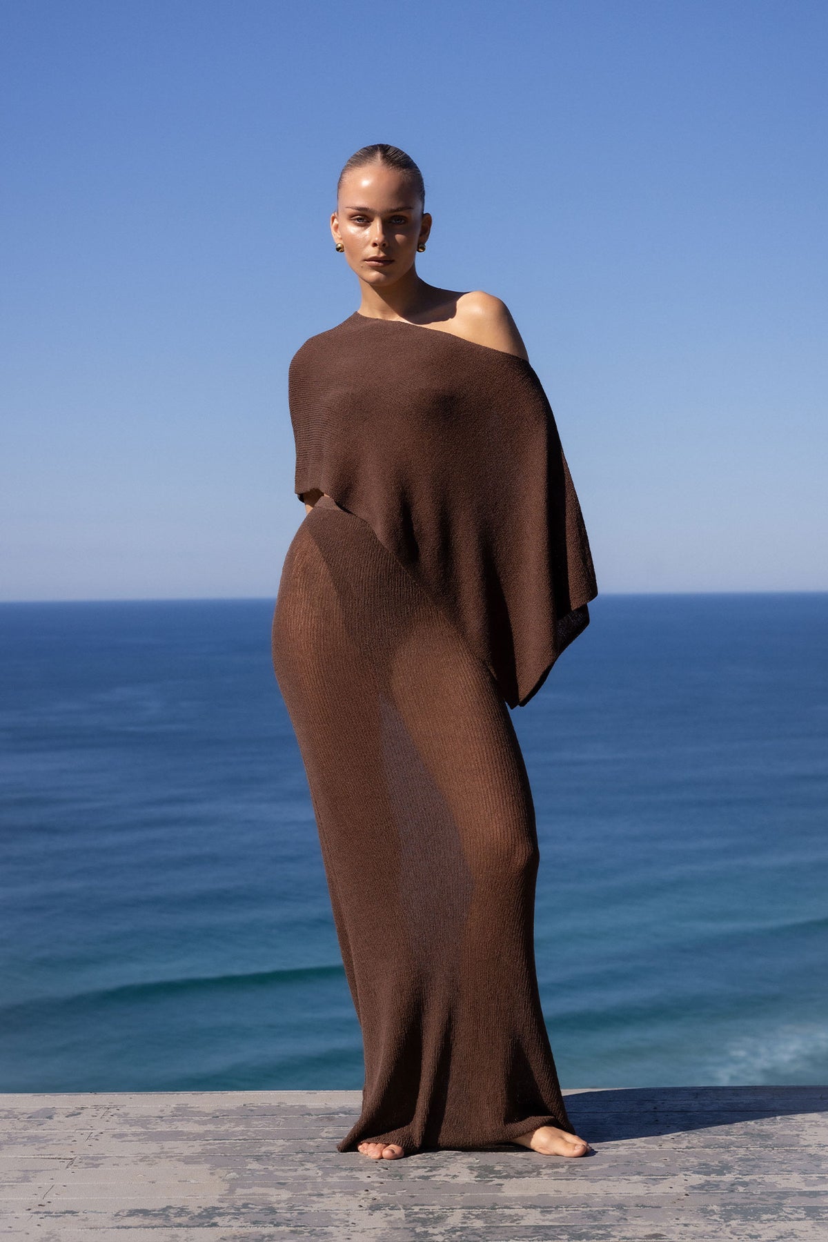 A model stands barefoot on a stone ledge overlooking the ocean, wrapped in a draped, one-shoulder brown knit cover-up or dress.