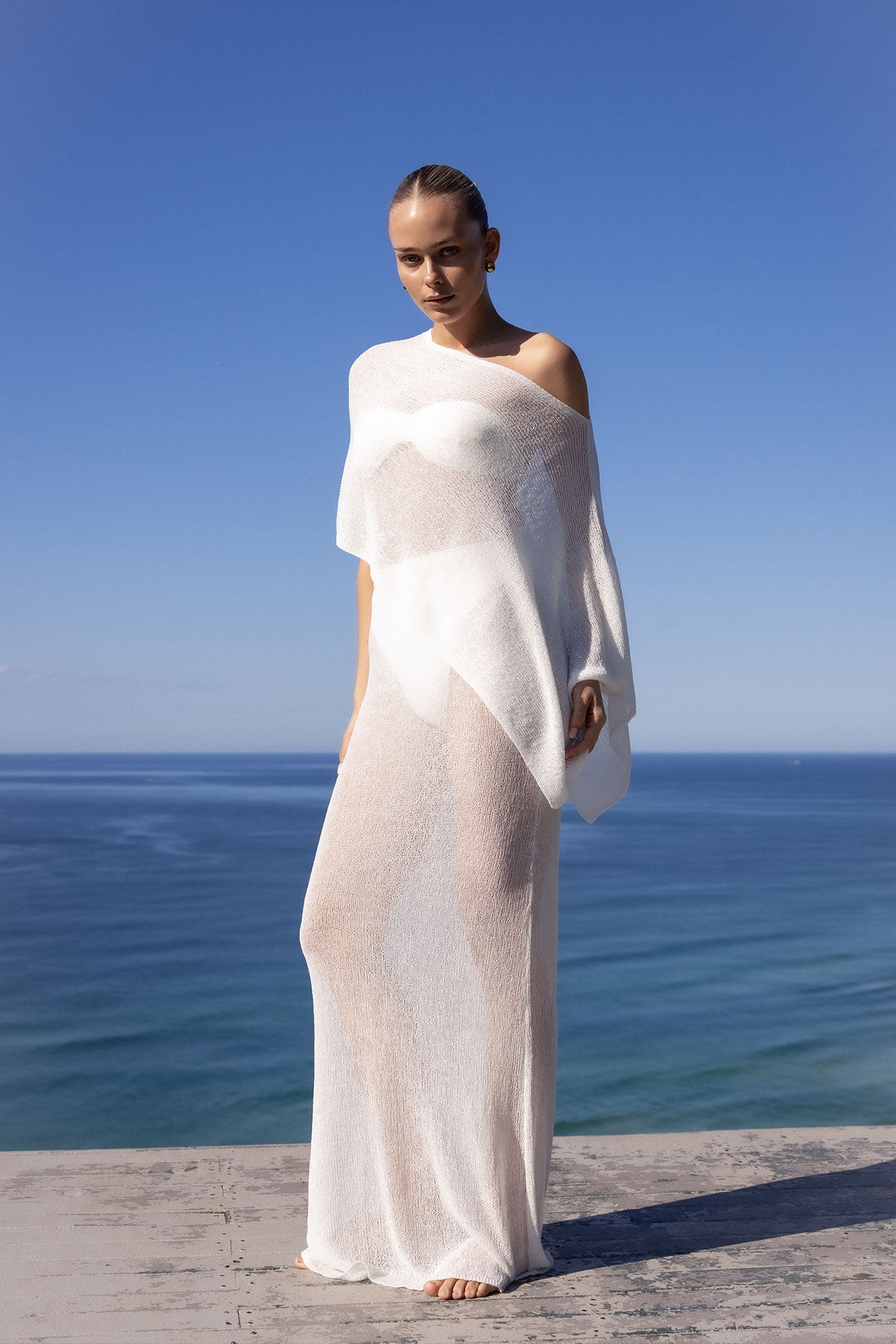 A woman stands barefoot on a stone ledge overlooking the ocean, wearing a sheer, off-the-shoulder white maxi dress or cover-up against a clear blue sky and sea background.