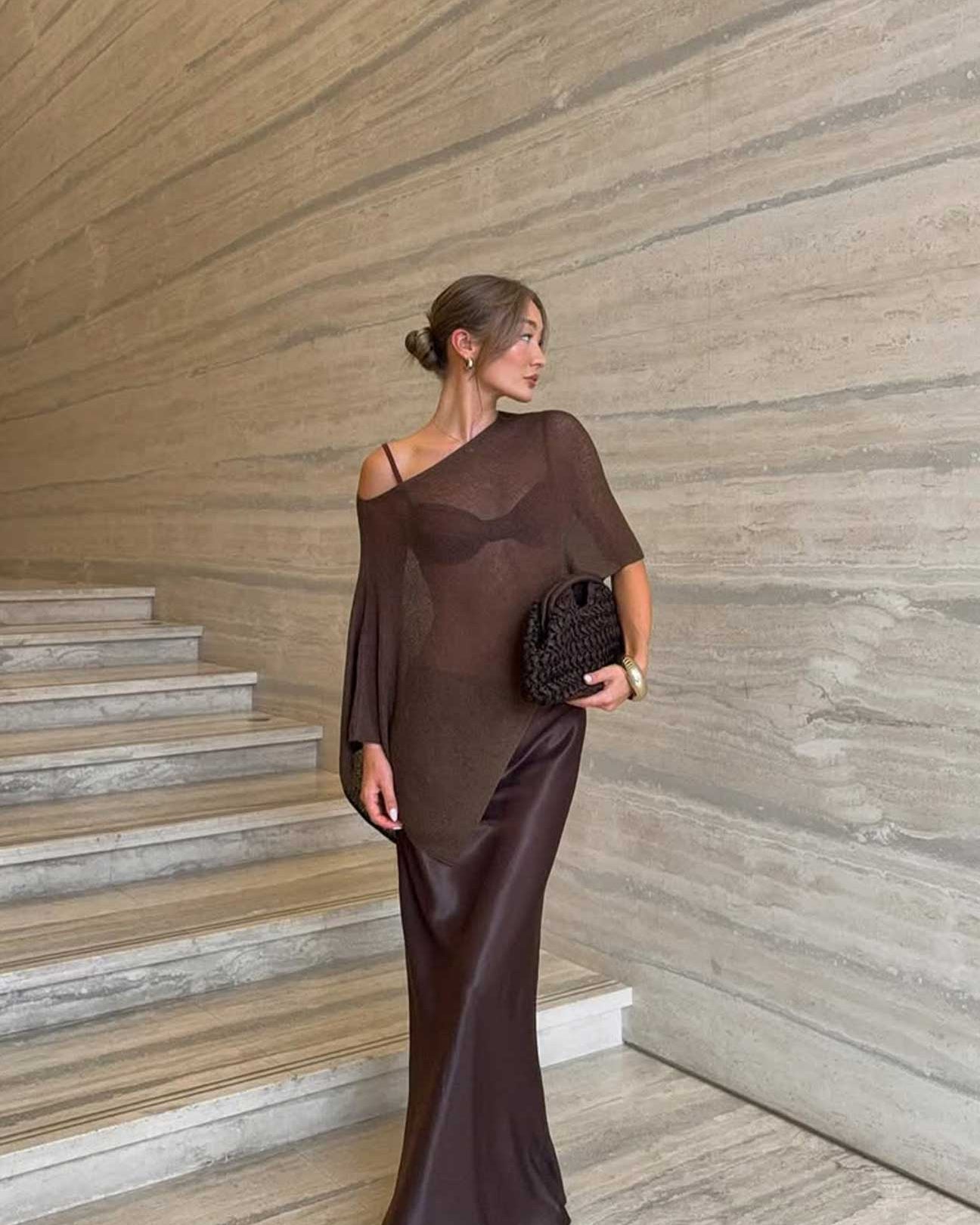 Woman in a dark brown off-shoulder cape standing on a staircase with a textured wall background