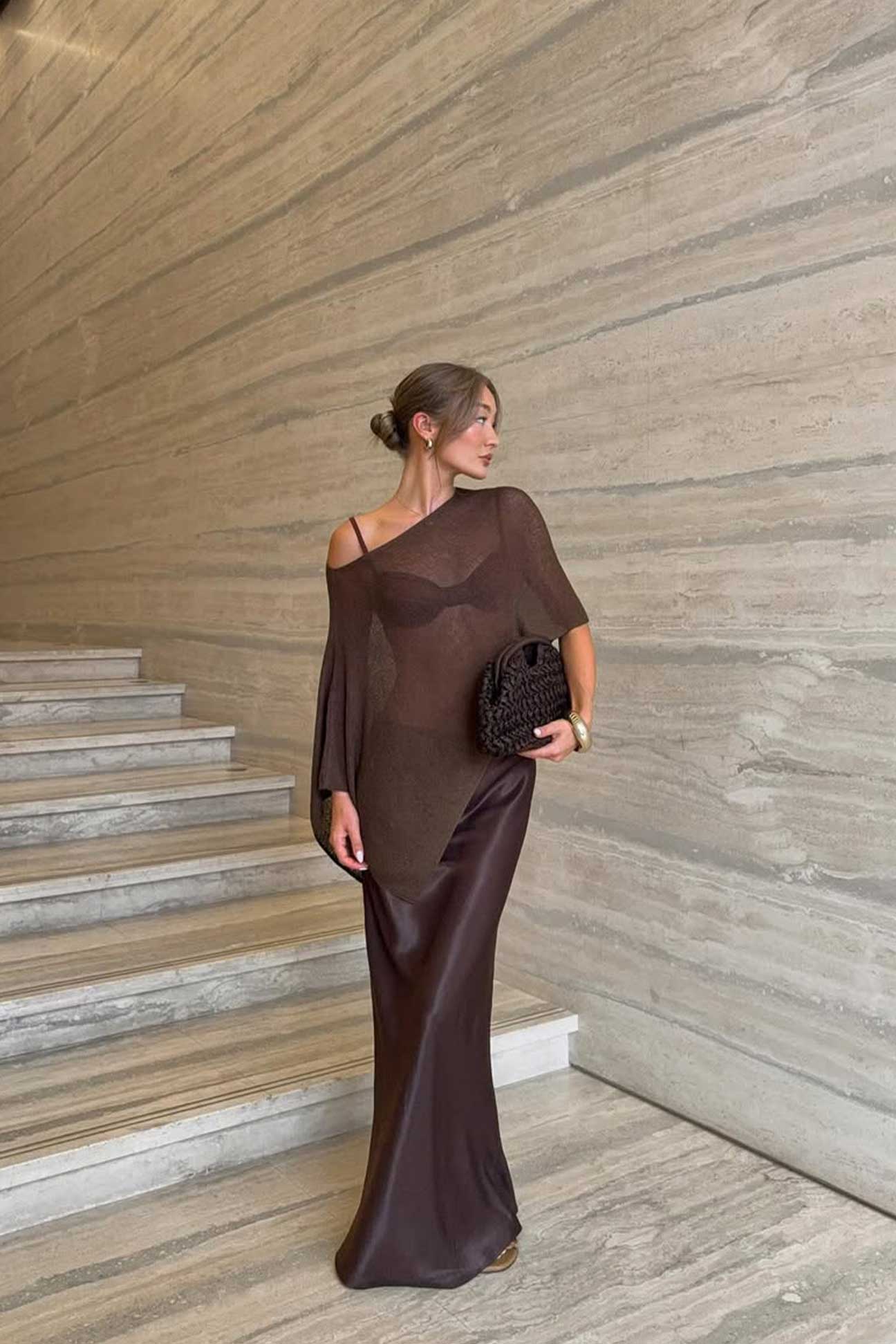 Woman in a dark brown off-shoulder cape standing on a staircase with a textured wall background