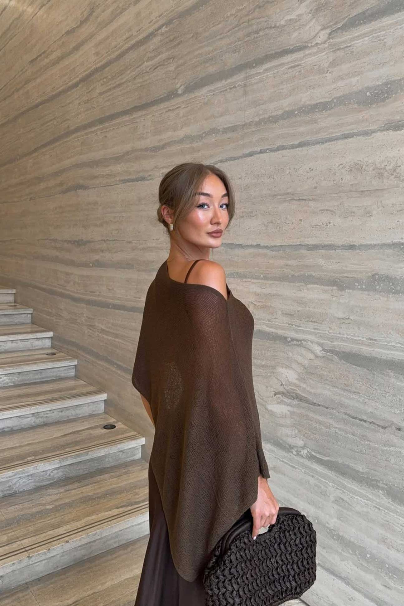 Woman in a brown cape standing on a staircase with a textured wall background
