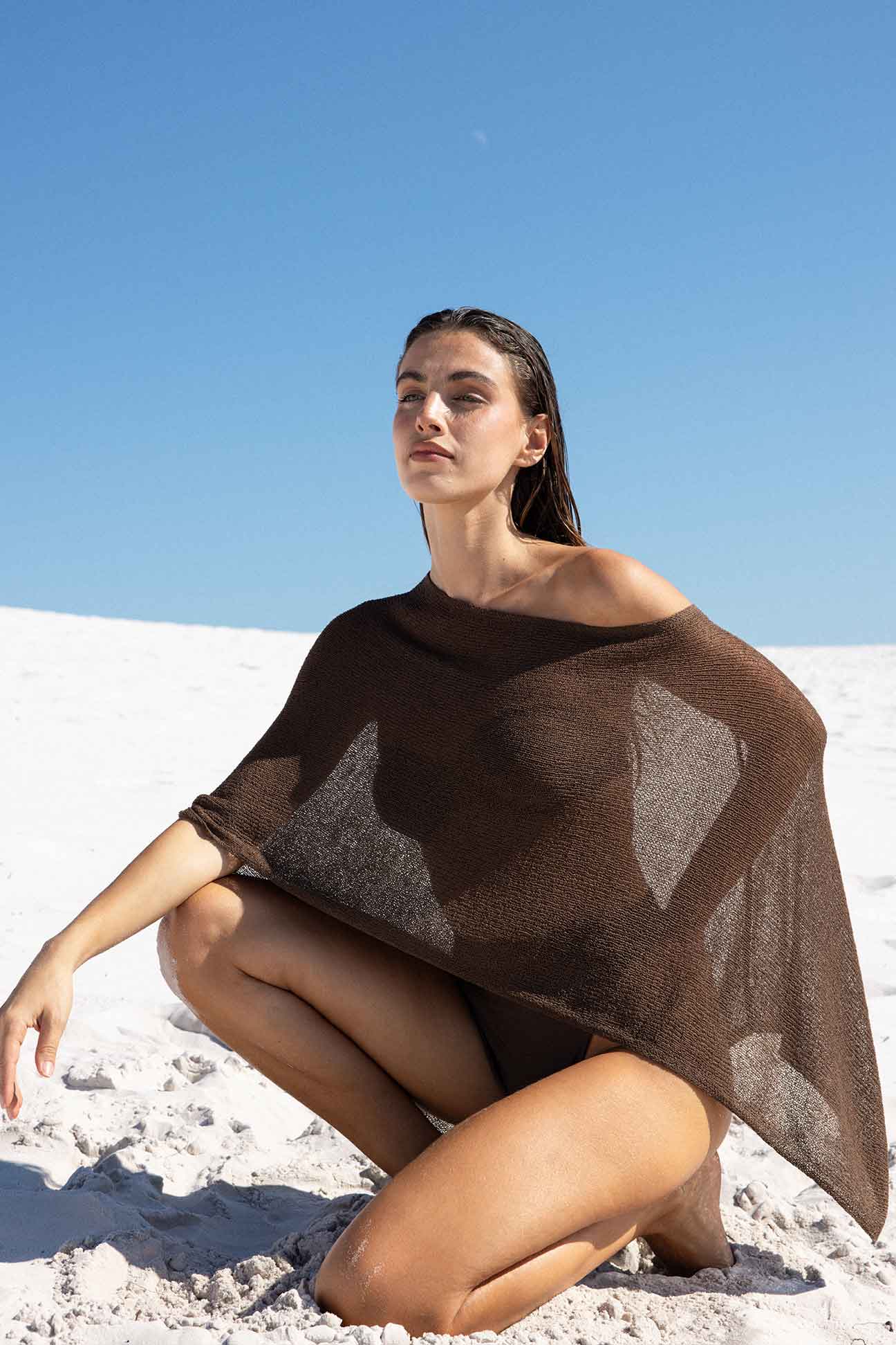 Woman sitting on a sandy surface wearing a brown poncho with a clear blue sky in the background
