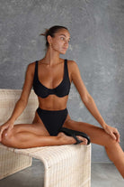 Woman in a black bikini sitting on a wicker stool against a gray wall.