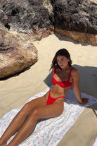 Woman in a red bikini sitting on a towel under large rocks on a sandy beach.