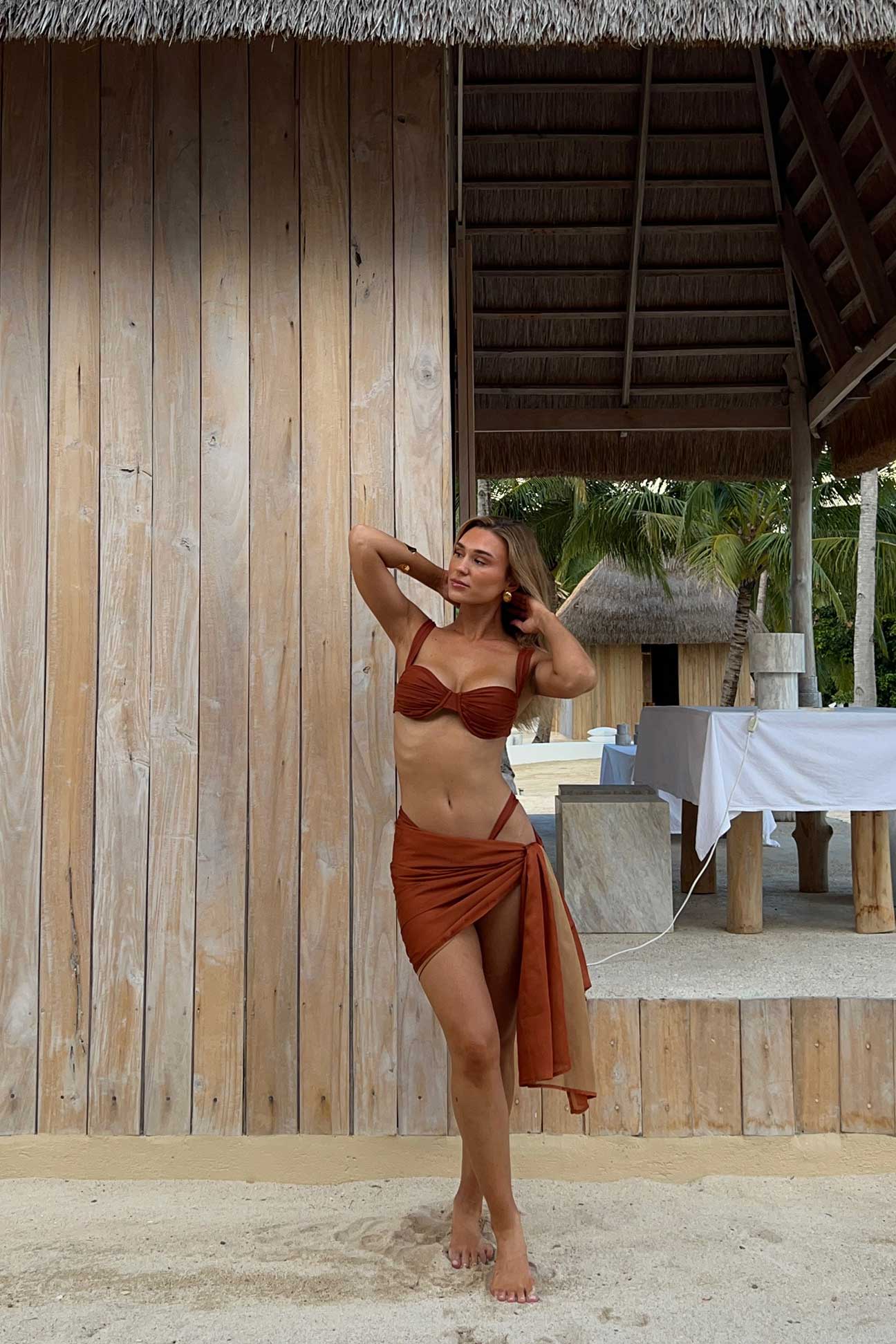 Woman in a terracotta bikini and sarong standing against a wooden wall with a tropical setting.