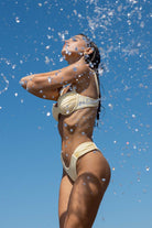 Woman in a yellow bikini standing under water splashes against a clear blue sky.
