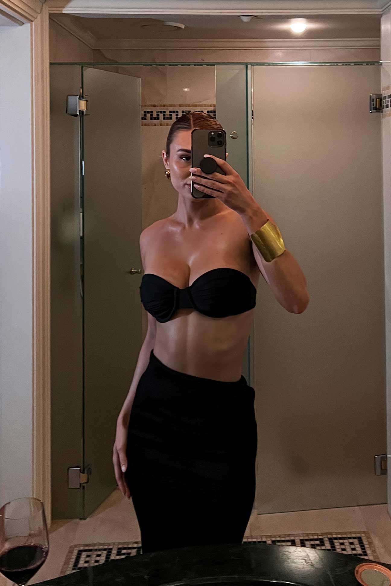 Person taking a mirror selfie wearing a black strapless bikini top and skirt in a bathroom.
