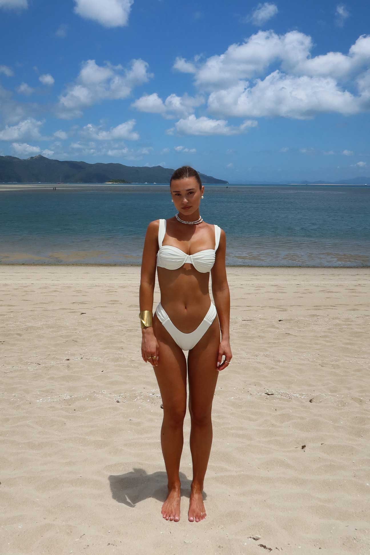 Woman in a white bikini standing on a sandy beach with a blue sky and mountains in the background.
