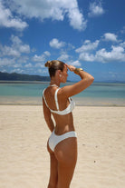 Woman in a white bikini standing on a sandy beach with blue sky and water in the background