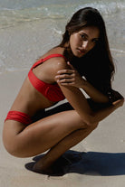 Woman in a red bikini squatting on a sandy beach.