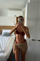 Person taking a mirror selfie in a bedroom wearing a terracotta bikini and beige wrap.
