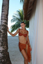 Woman in a terracotta bikini and sarong standing by a palm tree on a beach.