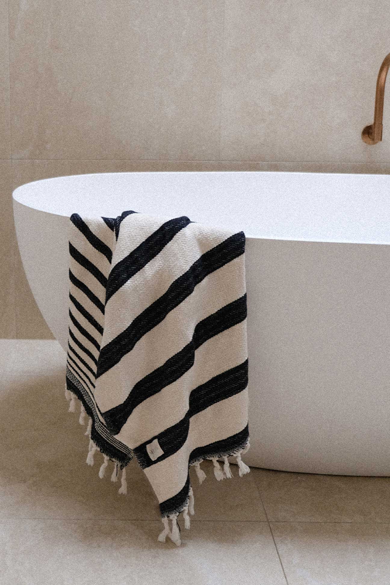 Black and white striped towel with tassels draped over a white bathtub in a bathroom setting.