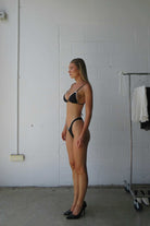 Woman wearing a black bikini standing against a white brick wall.