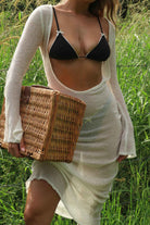 Woman in a black bikini top and white cover-up holding a woven basket in a grassy field