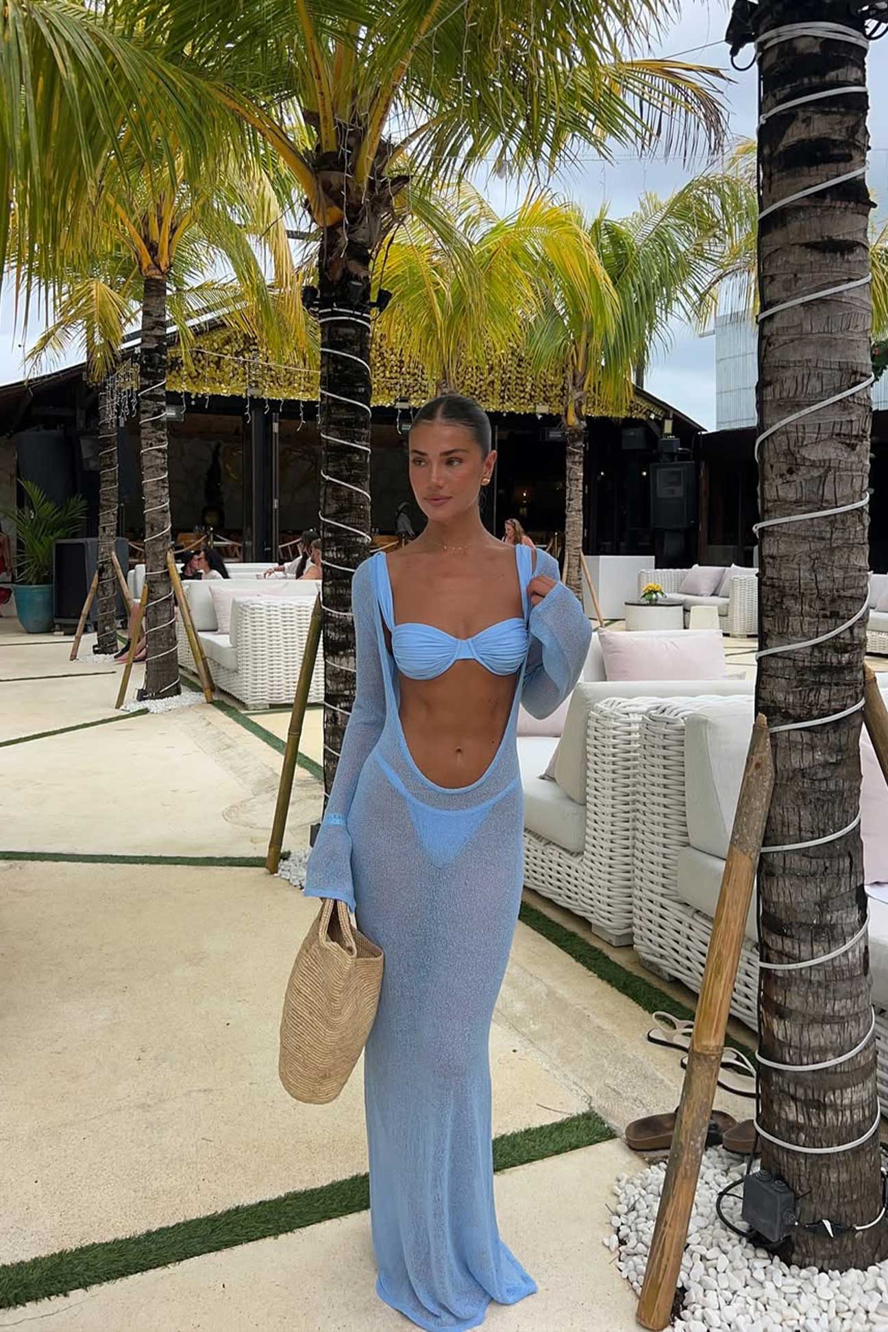 Woman in a blue dress standing in a tropical outdoor setting with palm trees and white furniture.